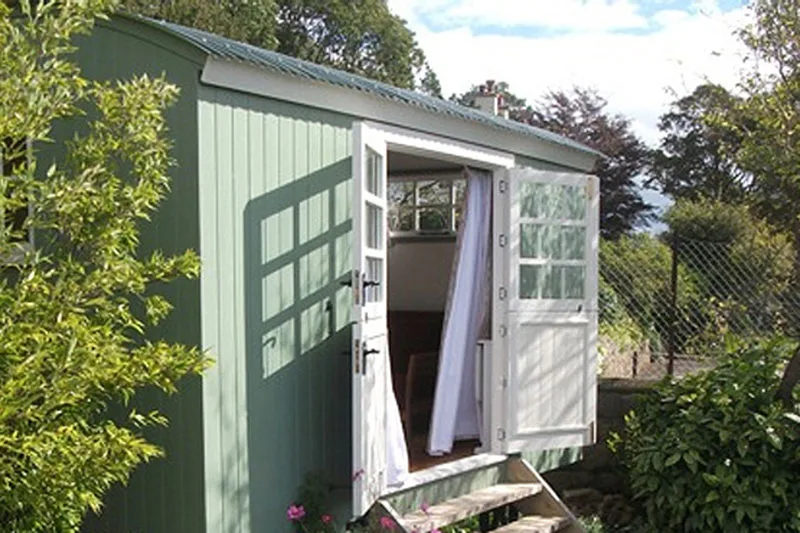 Light green hut nestled in the corner of a garden, double doors open letting the sun in.